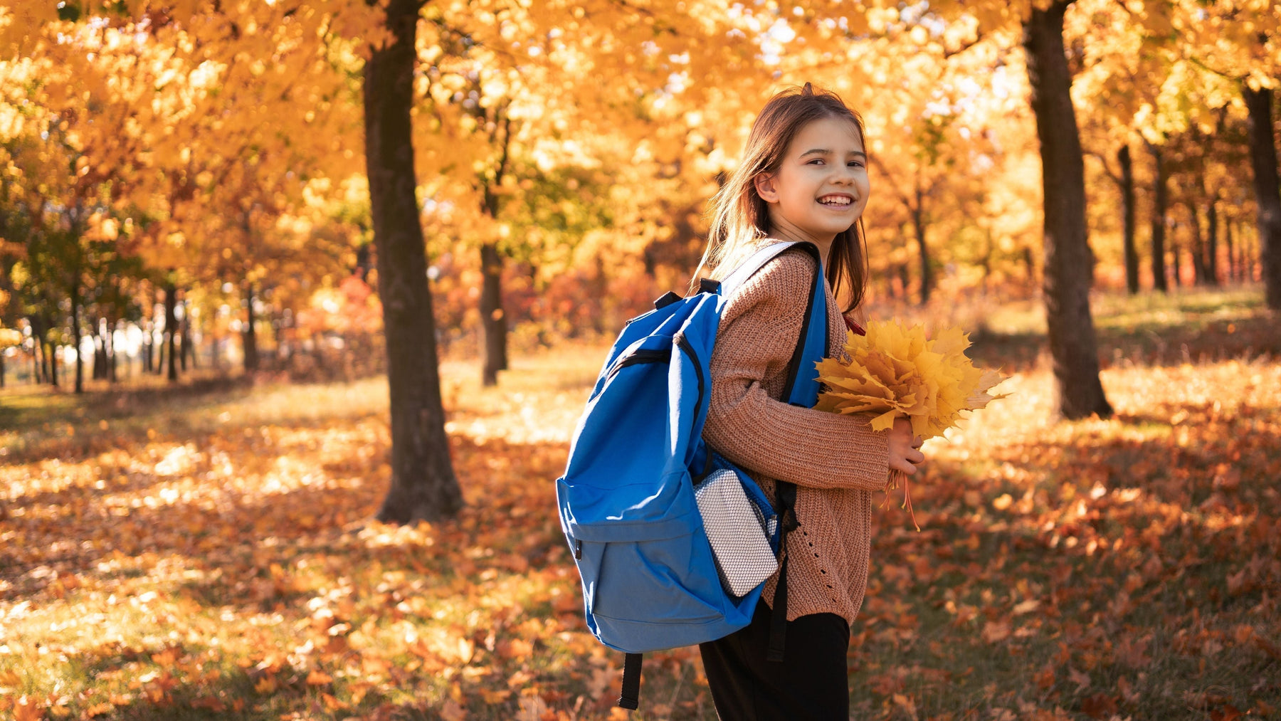 Smiling girl with blue backpack and autumn leaves in golden fall trees heading back to school