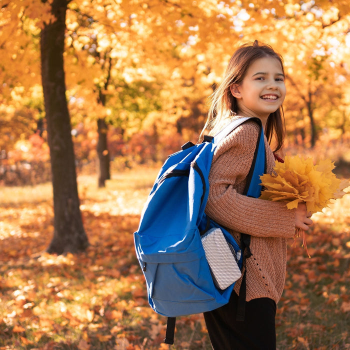 Smiling girl with blue backpack and autumn leaves in golden fall trees heading back to school