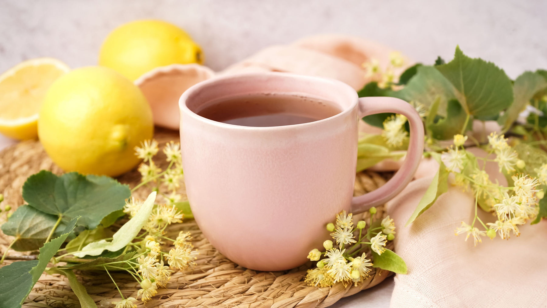 Pink mug of herbal tea with linden flowers lemon and fresh herbs on woven mat representing daily wellness habit