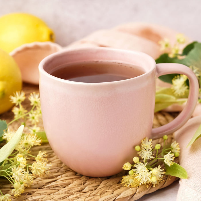 Pink mug of herbal tea with linden flowers lemon and fresh herbs on woven mat representing daily wellness habit