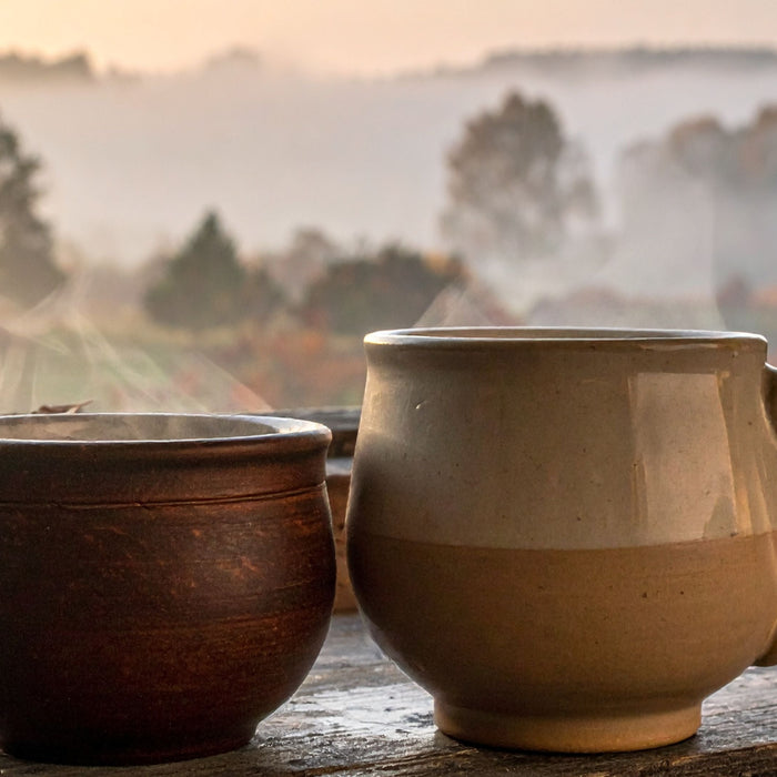 Two stoneware mugs of steaming tea on a wooden windowsill with a misty autumn landscape behind them