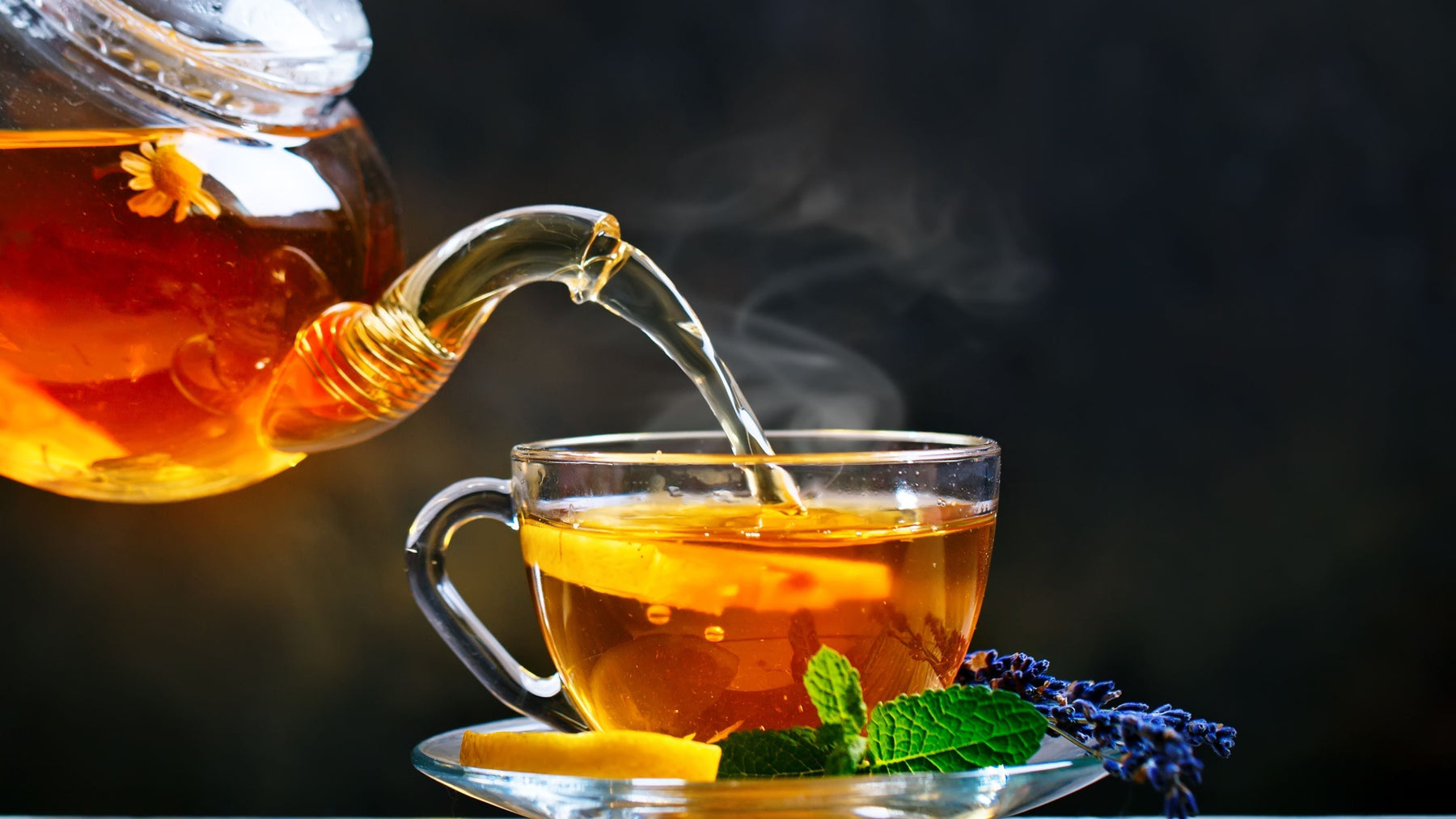 Glass teapot pouring steaming herbal tea into cup with lemon mint and lavender showing heat and brewing process