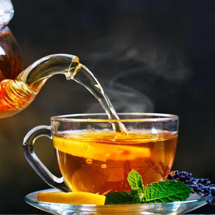 Glass teapot pouring steaming herbal tea into cup with lemon mint and lavender showing heat and brewing process