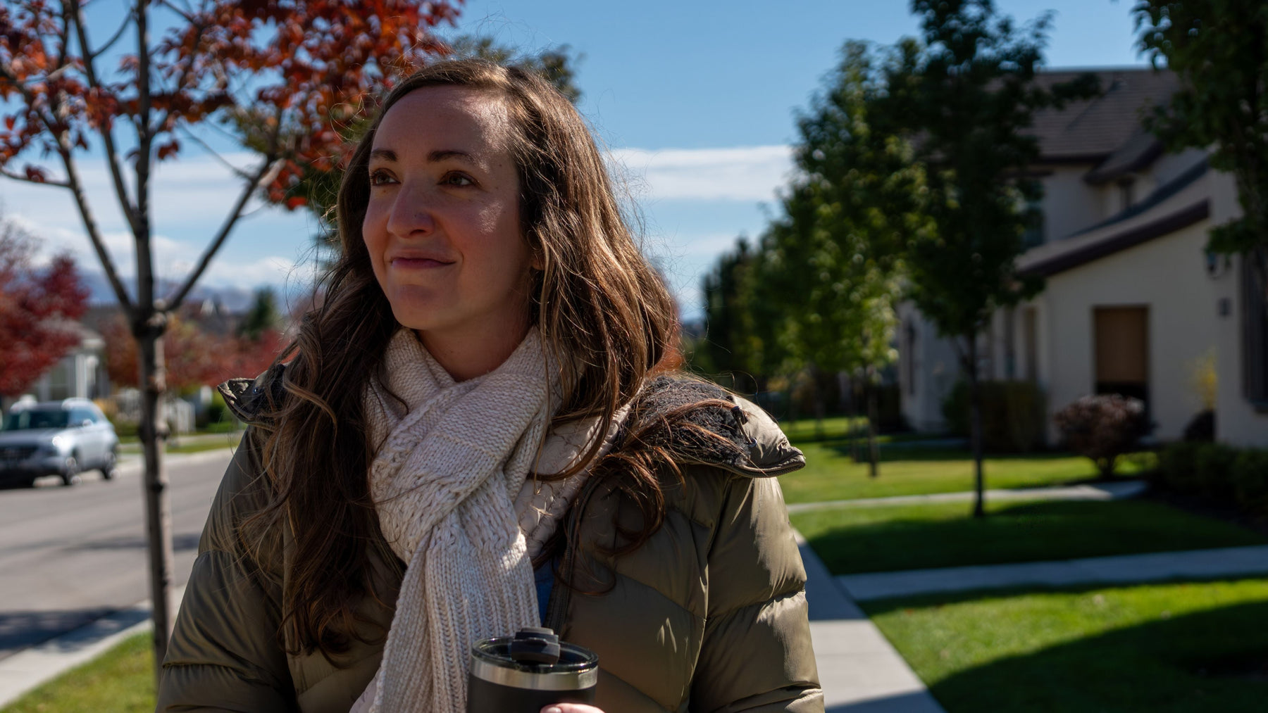 Smiling woman in a scarf and puffer jacket holding a travel tumbler on an autumn neighborhood walk