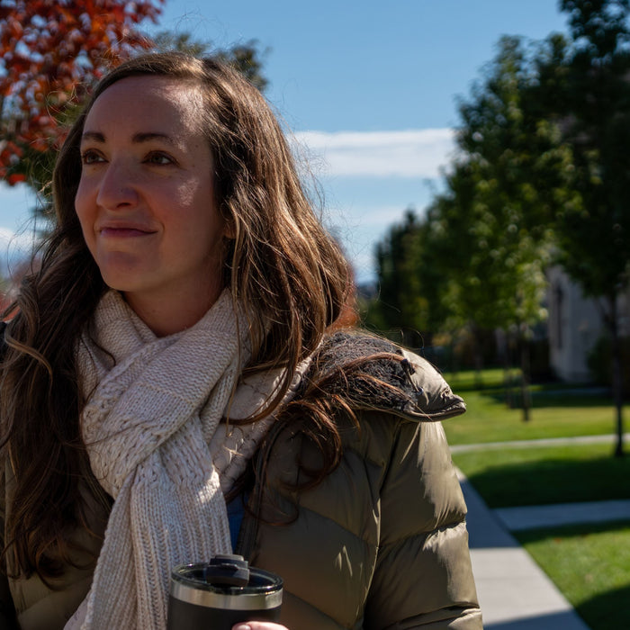 Smiling woman in a scarf and puffer jacket holding a travel tumbler on an autumn neighborhood walk