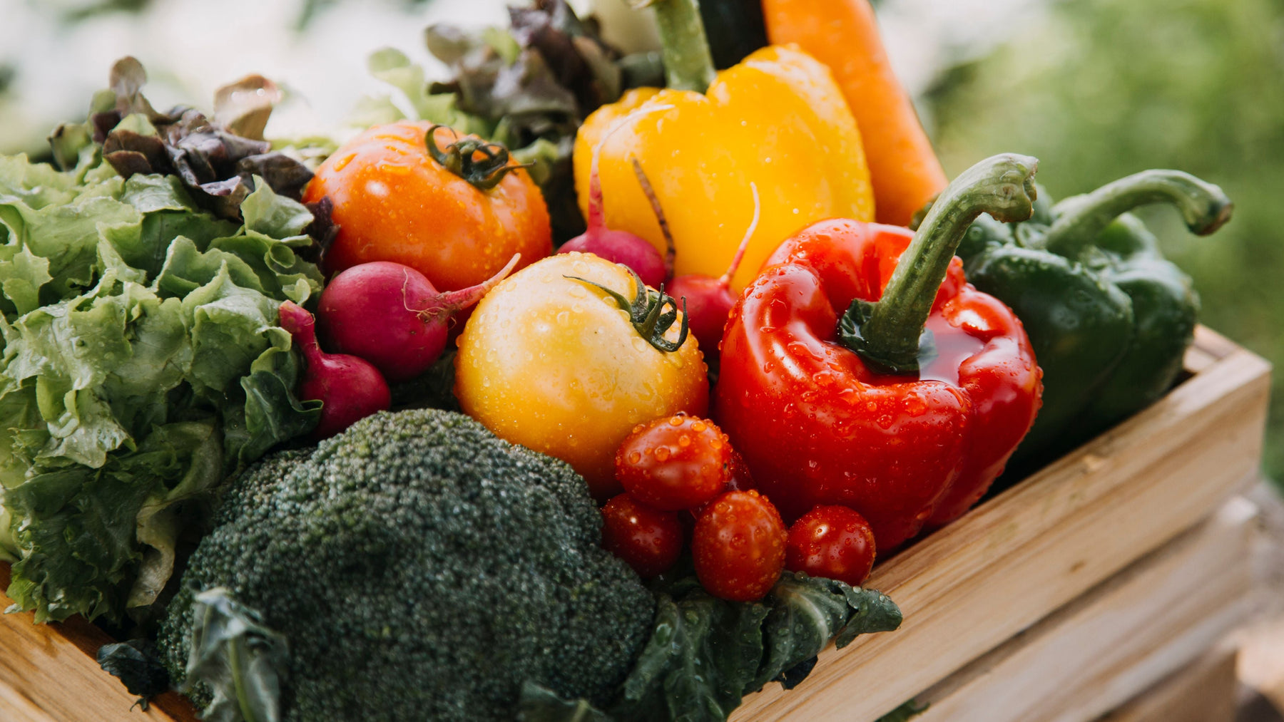 Wooden crate of fresh vegetables including broccoli, tomatoes, bell peppers, radishes, and lettuce