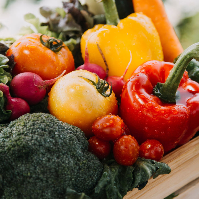Wooden crate of fresh vegetables including broccoli, tomatoes, bell peppers, radishes, and lettuce