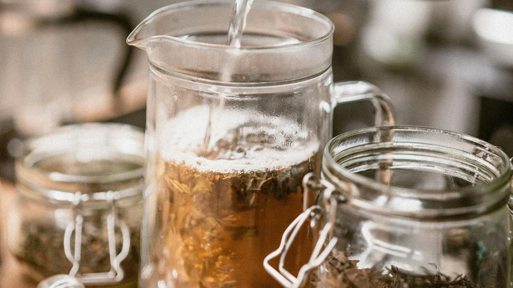 Water being poured over loose dried herbs in a glass pitcher, with jars of herbs on the counter