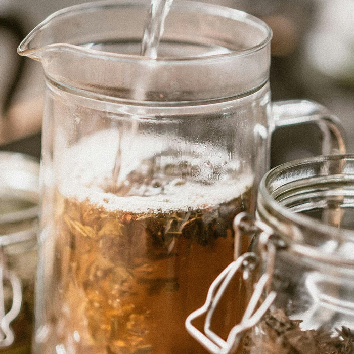 Water being poured over loose dried herbs in a glass pitcher, with jars of herbs on the counter