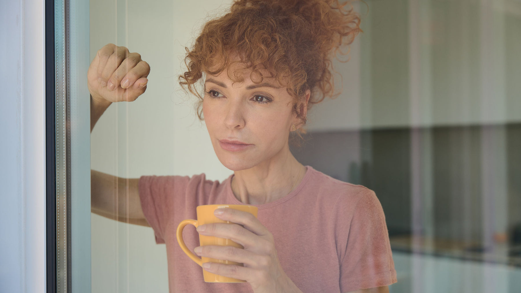 Thoughtful woman in a pink t-shirt holding an orange mug beside a window