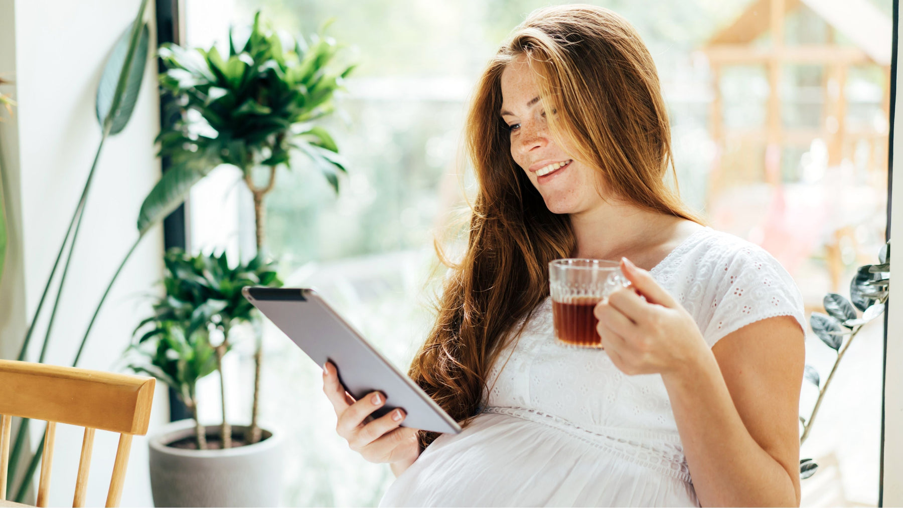 Pregnant woman smiling and drinking herbal tea while reading indoors with natural light and plants