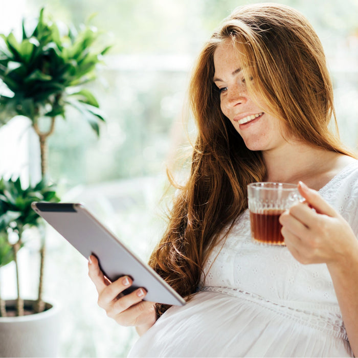 Pregnant woman smiling and drinking herbal tea while reading indoors with natural light and plants