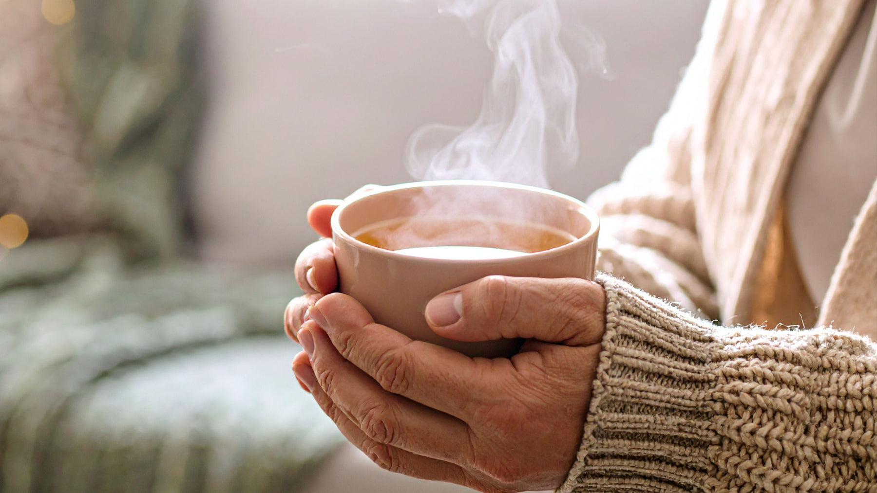 Hands in a cozy knit sweater holding a steaming mug of herbal tea