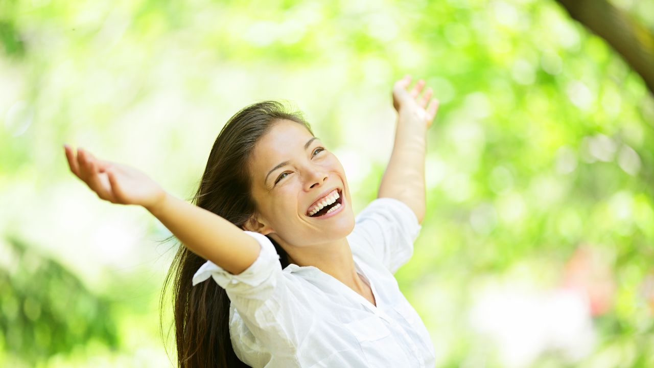 Woman outdoors in summer sunshine with arms raised among green trees